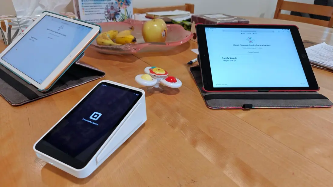 A staff member helping a parent check in on a tablet at the front desk, with the Square terminal beside it